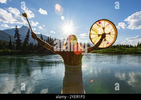 Una vista ravvicinata e posteriore di una donna sciamana che tiene un tamburo sacro della polvere verso il sole e le montagne durante il rituale di culto e guarigione, con spazio di copia Foto Stock