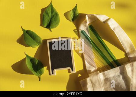 Cannucce in vetro riutilizzabili su sfondo colorato con foglietti verdi in sacchetto ecologico. Modello di simulazione della lavagna vuota per il testo dello spazio di copia. Bere Foto Stock