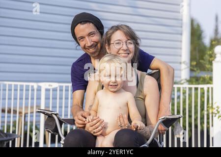 La giovane famiglia si mette in posa per un ritratto di gruppo visto dal davanti. Madre seduta sulla sedia con due anni di ragazzo in grembo e padre sorridente in piedi dietro Foto Stock