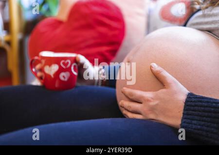 Una vista selettiva di fuoco di una donna gravida pesante con grande tummy gonfio rilassante all'interno del giorno delle valentines, con tazza blurry di tè in background Foto Stock