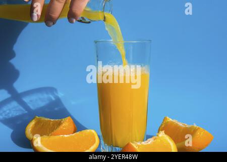 Versare il flusso di succo d'arancia dalla brocca in un bicchiere di succo d'arancia spremuto con frutta a fette su sfondo blu. Succo di frutta, bevanda fresca fredda. Rif Foto Stock