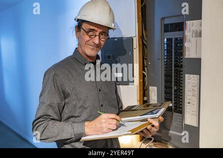 Ispettore domestico di fronte al quadro di distribuzione elettrica durante un'ispezione, un uomo caucasico che indossa casco e occhiali che prende appunti sul suo notebook Foto Stock