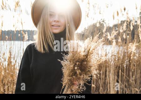 Donna bionda europea con cappello beige in pullover nero in campagna. Ora d'oro, cotagecore. Viaggi locali. Vita lenta. Salute mentale. Messa a terra a. Foto Stock