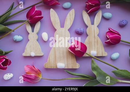 Uova di Pasqua e coniglietti di legno con tulipani rosa su sfondo viola pastello. Composizione piatta, vista dall'alto. Decorazioni dal design moderno minimalista Foto Stock