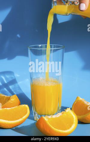 Versare il flusso di succo d'arancia dalla brocca in un bicchiere di succo d'arancia spremuto con frutta a fette su sfondo blu. Succo di frutta, bevanda fresca fredda. Rif Foto Stock