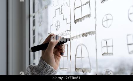 La mano femminile disegna le decorazioni natalizie dipinte su vetro della finestra. Paesaggi di New Years. Edifici della città ghirlande luci. Igge, decorazione e chri Foto Stock