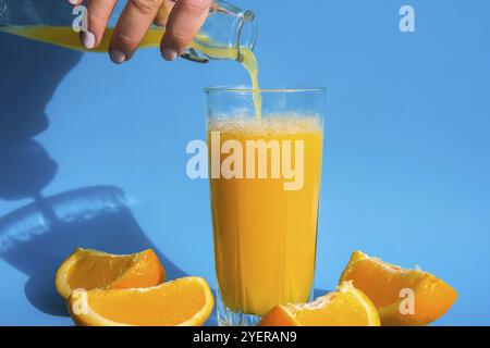 Versare il flusso di succo d'arancia dalla brocca in un bicchiere di succo d'arancia spremuto con frutta a fette su sfondo blu. Succo di frutta, bevanda fresca fredda. Rif Foto Stock