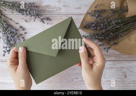 Composizione piatta di mani femminili che tengono una busta verde con fiori asciutti di lavanda. Copia modello spazio. Vista dall'alto. Concetto di invit di matrimonio Foto Stock