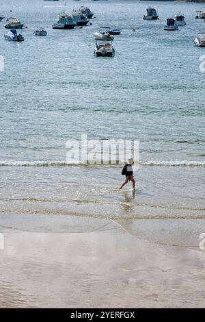 Donna che cammina lungo la spiaggia di la Concha a San Sebastian, in Spagna, con molte barche a vela in acqua Foto Stock