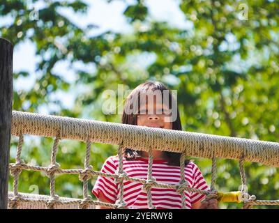 I bambini giocano all'aperto in un parco giochi in legno. Ragazza felice che gioca su un ponte di rete in corda nel parco giochi durante l'estate. Foto Stock