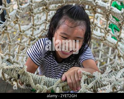 I bambini giocano all'aperto in un parco giochi in legno. Ragazza felice che gioca su un ponte di rete in corda nel parco giochi durante l'estate. Foto Stock