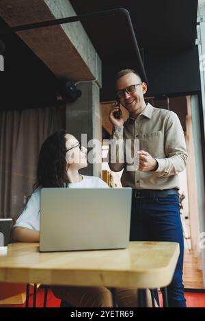 Un giovane uomo soddisfatto parla con una collega e si rallegra del lavoro svolto. I colleghi lavorano al computer portatile in ufficio. Lavorare in un ufficio moderno sp Foto Stock