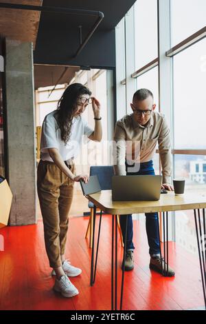 Un giovane uomo soddisfatto parla con una collega e si rallegra del lavoro svolto. I colleghi lavorano al computer portatile in ufficio. Lavorare in un ufficio moderno sp Foto Stock