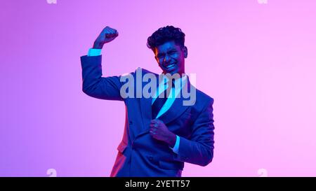 Ritratto di un giovane e allegro uomo indiano in abito formale con emozioni felici, alzando le mani e celebrando il successo su sfondo rosa in studio al neon Foto Stock
