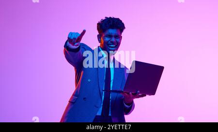 Ritratto di un uomo indiano felice, eccitato e sorridente in abito formale, con laptop e gesti, che mostra il successo sullo sfondo viola dello studio al neon Foto Stock