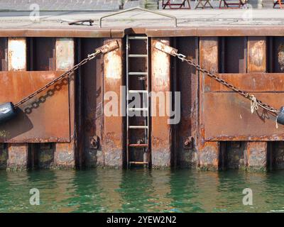 Parafango della banchina - sulla parete della banchina - il verde brillante dell'acqua contrasta nettamente con l'arrugginito palissandro, le catene e i parafanghi neri della banchina Foto Stock
