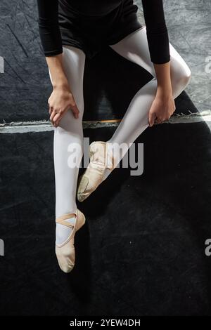 Ripresa verticale di una ballerina irriconoscibile che indossa pantofole di balletto che riposa dopo la lezione mentre si siede sul pavimento in uno studio di danza Foto Stock