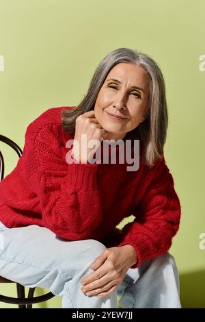 Un'elegante donna dai capelli grigi sorride in un comodo maglione rosso su uno sfondo verde brillante. Foto Stock