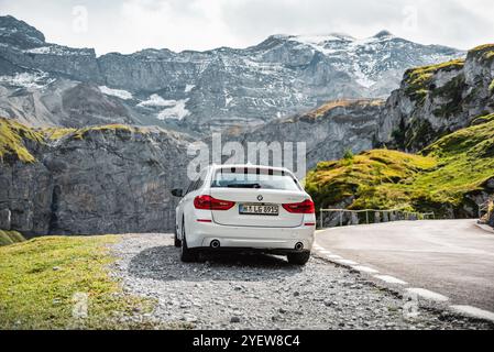 BMW serie 5 bianca parcheggiata su un lato di una strada tortuosa nelle alpi svizzere. Vista posteriore dell'auto tedesca, della scogliera e delle cime innevate sul retro. Foto Stock