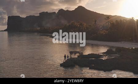 Due turisti in piedi sulla costa rocciosa, si godono il tramonto mozzafiato sulla silhouette di montagna a Maupiti, palme che costeggiano la spiaggia di sabbia bianca. La gente ama viaggiare in vacanza in una natura selvaggia e remota Foto Stock