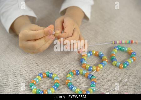 Primo piano delle mani di un bambino che infilano perline colorate su una corda, concentrandosi sulla creazione di un bracciale fatto a mano. Foto Stock