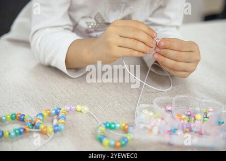 Primo piano delle mani di un bambino che infilano perline colorate su una corda, concentrandosi sulla creazione di un bracciale fatto a mano. Foto Stock