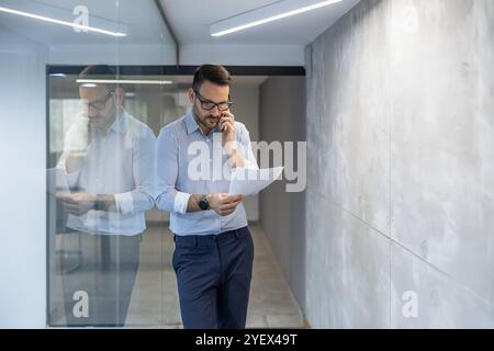 Un uomo d'affari professionista attraversa un moderno corridoio di uffici, impegnato in una conversazione telefonica durante la revisione di documenti importanti. La scena conv Foto Stock