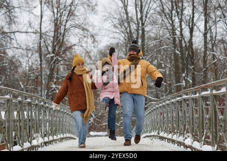 Ritratto completo di una famiglia felice con una figlia che salta e si tiene per mano mentre cammina verso la fotocamera sul ponte in inverno Foto Stock