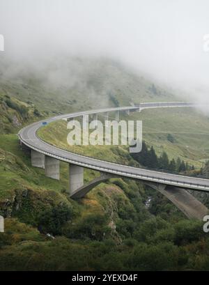 Pass del San Gottardo vuoto in una giornata nebbiosa. Grande curva di autostrada, pendio verde di montagna ricoperto di nebbia sullo sfondo. Strada nelle alpi svizzere. Foto Stock