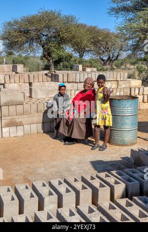 nonna africana del villaggio e bambini seduti su una sedia nel cortile tra un mucchio di mattoni Foto Stock