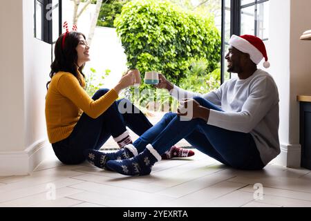 Festeggiamo il Natale, una coppia multirazziale con cappelli festivi che si gusta una bevanda calda a casa Foto Stock