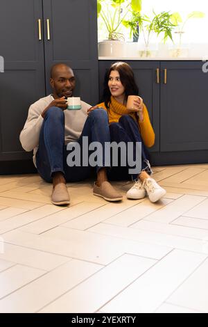 coppia multirazziale seduta sul pavimento della cucina, gustando un caffè e sorridendo insieme, a casa Foto Stock