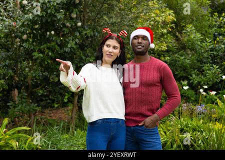 Coppia multirazziale in cappelli festivi che si godono il Natale all'aperto, puntano e sorridono Foto Stock