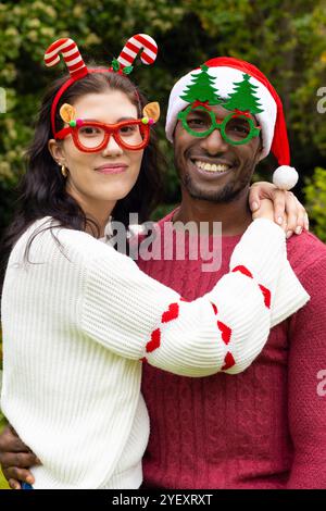 Coppia multirazziale sorridente che indossa occhiali e maglioni per festeggiare il Natale all'aperto Foto Stock