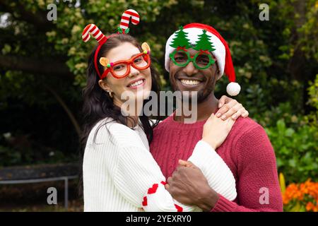 Coppia multirazziale sorridente che indossa occhiali e maglioni per festeggiare il Natale all'aperto Foto Stock