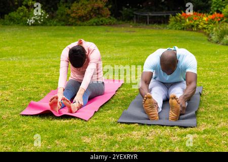 coppie multirazziali che praticano yoga su tappeti in giardino, si allungano e si rilassano all'aperto Foto Stock