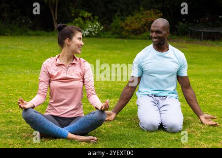 coppia multirazziale che pratica yoga insieme sull'erba, sorridendo e godendo del relax all'aperto Foto Stock