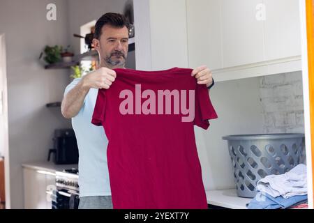 camicia rossa pieghevole uomo anziano in cucina con cesto per lavanderia nelle vicinanze, a casa Foto Stock