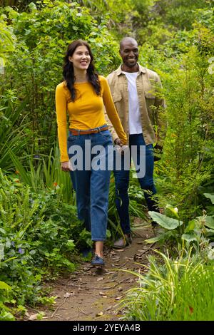 coppia multirazziale che cammina mano nella mano attraverso il lussureggiante giardino, godendosi la natura insieme Foto Stock