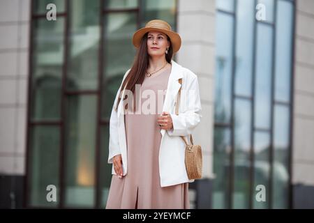 Elegante donna Plus size in piedi all'aperto in città, con abito beige, blazer bianco, cappello a teglia larga, borsa in tessuto. Modello con hai marrone lungo Foto Stock