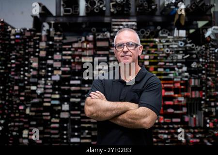 Operaio dell'industria metallurgica che posa con le braccia incrociate, dimostra competenza e orgoglio in un magazzino pieno di parti e attrezzature metalliche Foto Stock