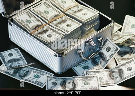 Una cassa metallica piena di pile di banconote da 100 dollari, isolata su uno sfondo scuro, con banconote sciolte sul tavolo Foto Stock