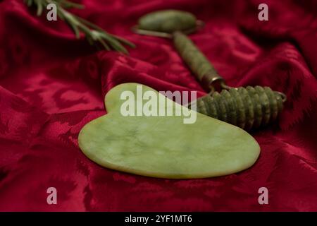 Pietra e rullo per massaggio viso gua Sha giada verde su sfondo rosso seta, primo piano Foto Stock