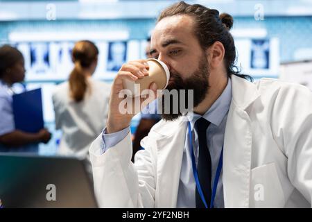Preoccupato medico stanco che beve caffè mentre esamina i risultati dei test, alla ricerca di diagnosi sul notebook. Medico in stress che legge i registri ospedalieri e altri rapporti per la prevenzione delle malattie. Foto Stock