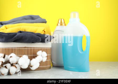 Detersivo per bucato in bottiglie, fiori di cotone e vestiti su un tavolo in marmo grigio su sfondo giallo Foto Stock