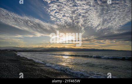 Uno splendido tramonto proietta sfumature dorate attraverso le dolci onde che si snodano su una spiaggia di ciottoli, incorniciata da nuvole vibranti. Foto Stock