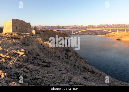 Donna turistica in piedi su una roccia al tramonto presso le rovine di un'antica fortezza Fortica sull'isola di pag, Dalmazia, Croazia, ponte pag sul retro Foto Stock