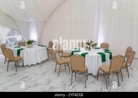 Bicchieri da vino, stoviglie in argento e tovaglioli verdi su tavoli rotondi ricoperti di tovaglia bianca. Concetto di catering. Interno della tenda per la cena di nozze, Foto Stock