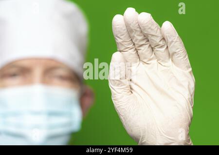 Medico maschile in chirurgia maschera viso su sfocatura primo piano mettere in avanti palmo della mano in guanti medici protettivi in primo piano, onde la sua mano, stop o lui Foto Stock