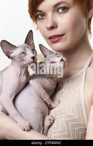 Cute giovane donna rossa abbracciando delicatamente due Sphynx Cat domestici al suo petto e guardando la macchina fotografica. Fuoco selettivo sui gattini amichevoli senza capelli. Prigioniero Foto Stock